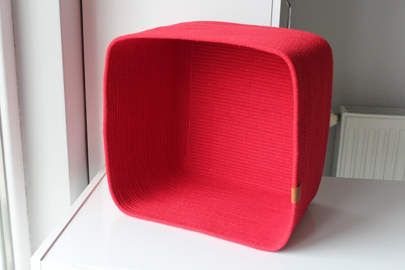 Puede incluir: Una cesta de almacenamiento cuadrada construida con cuerda roja enrollada. La cesta presenta esquinas redondeadas y una peque&ntilde;a leng&uuml;eta rectangular de color cuero tostado unida al lado inferior derecho de la pared exterior.