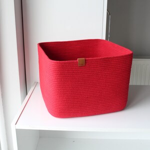 Puede incluir: Una cesta de almacenamiento cuadrada construida con cuerda enrollada de color rojo vibrante. La cesta presenta un dise&ntilde;o estructurado de parte superior abierta con esquinas redondeadas y una peque&ntilde;a etiqueta rectangular de color tostado.