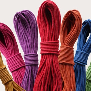 Op de afbeelding: Een verzameling van zeven spoelen met gekleurde koord, elk strak opgewonden en gepresenteerd tegen een witte achtergrond. De koorden zijn in tinten oranje, roze, paars, rood, bruin, blauw en groen.