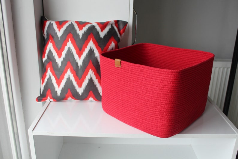 Puede incluir: Una cesta de almacenamiento cuadrada de cuerda roja con una peque&ntilde;a leng&uuml;eta de cuero marr&oacute;n en el borde superior, colocada junto a un coj&iacute;n cuadrado con un patr&oacute;n de zigzag en chevron rojo, blanco y gris oscuro sobre un estante blanco.