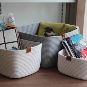 May include: Three white rope storage baskets with brown leather handles. The baskets are filled with fabric featuring bird and floral designs.
