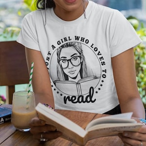 Op de afbeelding: Een wit T-shirt met een zwart-witte afbeelding van een meisje dat een boek leest. De afbeelding is omringd door de tekst "Just a girl who loves to read".