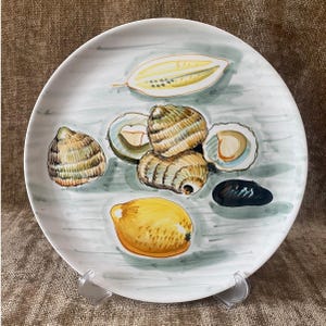 Puede incluir: Un plato de cerámica blanco con un diseño pintado a mano que presenta limones, ostras y almejas. El plato tiene un fondo azul claro y gris con una estética rústica. El plato se exhibe en un soporte de plástico transparente.