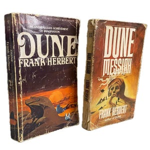 May include: Two vintage paperback books, "Dune" and "Dune Messiah" by Frank Herbert. The "Dune" cover features a desert landscape with the title in large white letters. "Dune Messiah" has an orange cover with a portrait and the title in white.