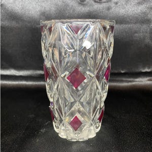 May include: A clear glass vase with a geometric diamond pattern and burgundy-colored diamond accents. The vase has a flared top and a textured, faceted design, suitable for displaying flowers or as a decorative piece.