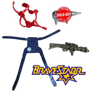 May include: A collection of vintage BraveStarr action figure accessories. Includes a red, organic-shaped piece, a silver drill with a "SOLD OUT" sticker, a dark blue harness with a star emblem, and a gray rifle. The BraveStarr logo is also visible.