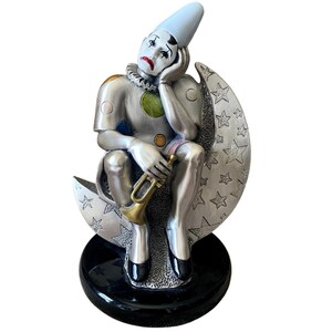 May include: A silver-toned clown figurine seated on a crescent moon, holding a trumpet. The clown wears a white hat, a ruffled collar, and a sad expression. The moon is decorated with stars and rests on a black base. A decorative item.