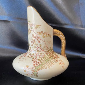 Puede incluir: Jarra de cerámica color crema con asa y borde dorados. La jarra está decorada con un diseño botánico de hojas y vides rojas, verdes y marrones. Tiene una base bulbosa y un pico curvo. Altura aproximada: 20 cm.
