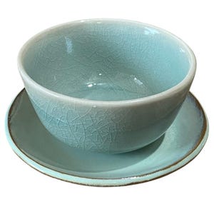 May include: A light blue ceramic bowl and saucer set. The bowl has a rounded shape with a textured, crackled surface. The saucer is circular with a slightly raised edge. The set has a glossy finish and a brown trim.