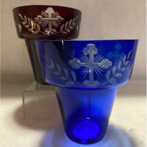 May include: Two glass candle holders, one deep red and one blue. Each has a cross design etched into the glass, surrounded by a leaf pattern. The blue candle holder is in the foreground.