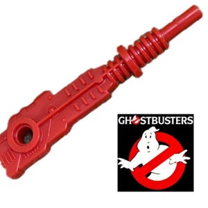 May include: A red plastic toy gun accessory with a detailed design. The Ghostbusters logo is visible in the lower right corner, featuring the iconic ghost symbol inside a red no-entry sign.