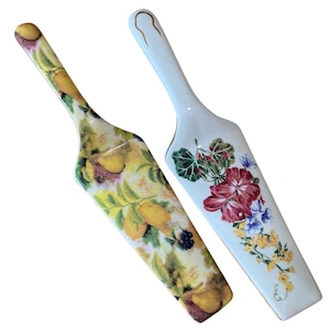 May include: Two decorative cake servers. One features a lemon and fruit design, the other has a floral pattern with red, purple, and yellow flowers. Both have long, white handles with gold accents.