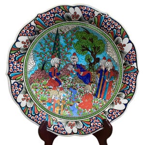 Puede incluir: Plato de cerámica decorativo con borde festoneado, que presenta una ilustración detallada y colorida de figuras en una escena de jardín. El plato tiene una base blanca con un borde floral en rojo, azul y verde.