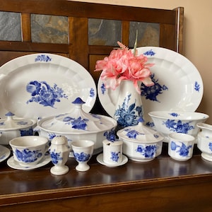 May include: A set of blue and white porcelain dinnerware featuring floral designs. The set includes a large platter, a serving bowl, a gravy boat, a sugar bowl, a creamer, and several smaller bowls. The dinnerware is displayed on a wooden surface with a bouquet of pink flowers.