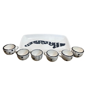 May include: A white ceramic serving tray featuring a black domino-inspired graphic, accompanied by six matching small cups. Each cup is decorated with black dots and lines, mirroring the domino theme found on the rectangular serving platter.