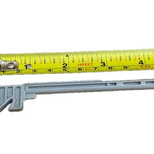 May include: A gray plastic tool with a handle and a long, thin shaft. The tool is about 5 inches long and has a measurement tape in the background.