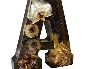 Könnte beinhalten: Ein Harzbuchstabe "A" gefüllt mit getrockneten Blumen in Weiß-, Creme- und Lilatönen. Der Buchstabe hat einen dunklen, glitzernden Hintergrund, und die Blumen sind im klaren Harz sichtbar.