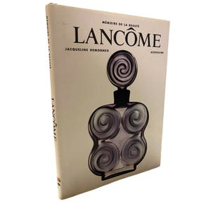 Puede incluir: Un libro titulado "Mémoire de la Beauté" de Lancôme, con una ilustración de un frasco de perfume. La portada es de color blanco roto con texto negro y un diseño de frasco de perfume morado y negro. También es visible el nombre de la autora, Jacqueline Demornex.