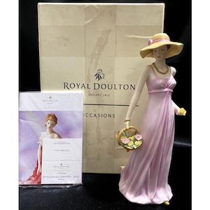 Op de afbeelding: Royal Doulton beeldje van een vrouw in een roze jurk en een gele hoed, met een mandje bloemen. Het beeldje wordt gepresenteerd met de originele doos en een kaart. Op de doos staat "Royal Doulton England 1815".