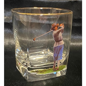 Könnte beinhalten: Ein durchsichtiges Glas mit einem goldfarbenen Rand und einer farbigen Grafik eines Golfers, der einen Hut, ein weißes Hemd und einen blauen Rock trägt und gerade mit einem Golfschläger ausholt. Der Boden des Glases zeigt eine grüne Grasfläche und ein kleines Wasserhindernis.