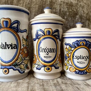 May include: Three white ceramic canisters with blue and yellow decorative borders and floral motifs. The tallest canister is labeled Salvia, the middle one is labeled Oregano, and the shortest one is labeled Espliego. Each features a blue ribbon design.