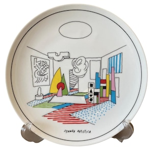 May include: White ceramic plate with a black border and a colourful abstract interior design. The design includes geometric shapes, lines, and the text "STANZA ARTISTICA". The plate is displayed on a clear stand.