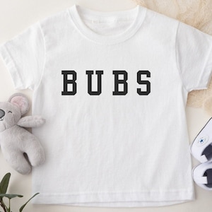 May include: White t-shirt with the word "BUBS" printed in black block letters.