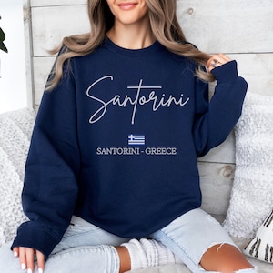 May include: Navy blue sweatshirt with white text that reads "Santorini" and a small Greek flag. The text "Santorini - Greece" is printed below the flag.