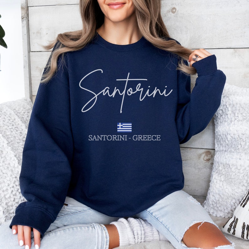May include: Navy blue sweatshirt with white text that reads "Santorini" and a small Greek flag. The text "Santorini - Greece" is printed below the flag.