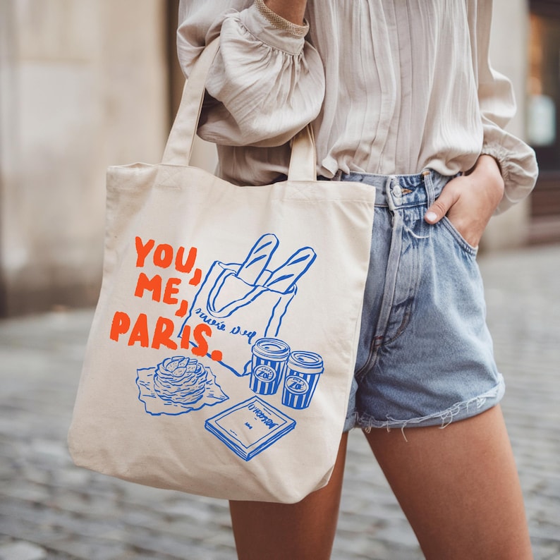 May include: A beige tote bag with the words "YOU, ME, PARIS" in orange. The bag features blue illustrations of baguettes, coffee cups, and a pastry. The bag is carried by a person wearing a light-colored blouse and denim shorts.