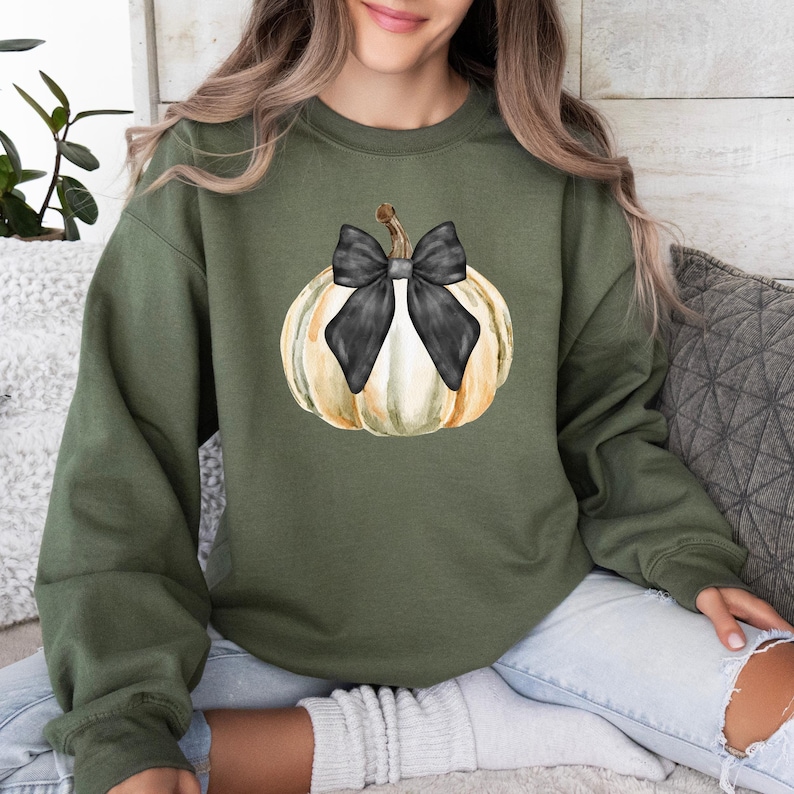 May include: Olive green sweatshirt featuring a watercolor pumpkin design. The pumpkin is white and yellow with a black bow. The sweatshirt is a comfortable, casual style, perfect for fall.