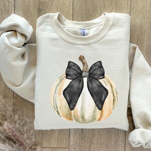 May include: Cream-colored sweatshirt featuring a watercolor pumpkin design with a black bow. The pumpkin has shades of white, green, and orange. The sweatshirt is folded on a wooden surface.