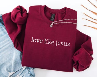 May include: A maroon crewneck sweatshirt with the text "love like jesus" in white. A silver necklace with a pendant is draped across the shirt. The sweatshirt is folded on top of a pair of blue jeans.