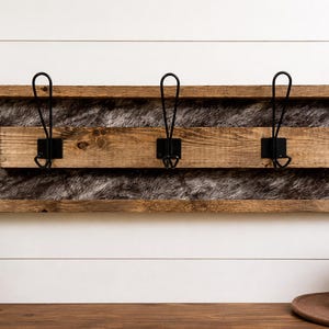 Könnte beinhalten: Ein rustikaler Wand-Kleiderhaken mit drei schwarzen Metallhaken. Der Kleiderhaken hat einen Holzrahmen mit einem Kunstfell-Einsatz. Die Holzbasis hat eine natürliche Oberfläche, und die Haken sind gleichmäßig verteilt.