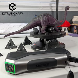 Puede incluir: Una figura de dinosaurio morada sobre una base giratoria negra con pequeños marcadores negros. En primer plano, un dispositivo MetroX plateado y negro con una luz verde brillante conectado por un cable negro, junto a varios marcadores triangulares negros pequeños.