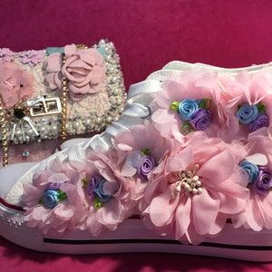 Może przedstawiać: Białe płócienne buty sportowe o wysokiej cholewce z różowymi i niebieskimi kwiatami z tkaniny. Buty mają białą gumową podeszwę i czarną obwódkę wokół krawędzi.