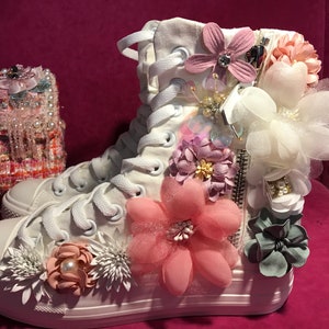 Może przedstawiać: Białe sneakersy wysokie z zamkiem błyskawicznym i ozdobione różowymi, białymi i jasnofioletowymi kwiatami z tkaniny. Sneakersy znajdują się na różowym tle.