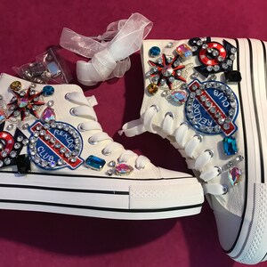 Może przedstawiać: Białe wysokie trampki ozdobione kolorowymi dżetami, naszywkami i wstążkami. Trampki mają czarno-białą, paskowaną podeszwę i napis "REKIDS CLUB" na naszywce.