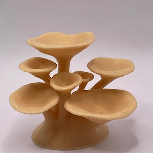 May include: A beige, mushroom-shaped decorative object. The object features multiple mushroom caps of varying sizes, supported by a central stem. The caps have a smooth, rounded shape, and the stem has a textured appearance. The object is set against a plain background.