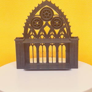 May include: A black, decorative architectural model featuring a gothic arch design. The model has three circular starburst patterns at the top and a series of vertical bars below. The model is set against a yellow background.