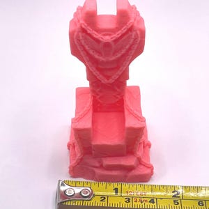 May include: A pink 3D-printed throne with intricate details, including a decorative chain around the top. The throne is approximately 3 inches tall, with a textured base. A yellow tape measure is placed next to the throne.