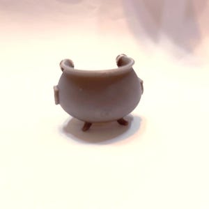 May include: A small gray cauldron with three legs and two handles. The cauldron is made of a smooth, matte material.