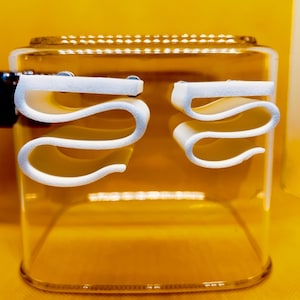 May include: A clear glass container with a gold-colored liquid inside. Two white, wavy, S-shaped holders are attached to the front. The container is on a yellow surface.
