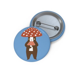 Può includere: Un bottone blu con un orso marrone che indossa un cappello di fungo rosso e bianco e tiene un fungo rosso e bianco.