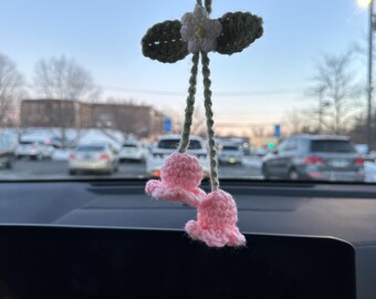 May include: A pink and green crocheted flower car charm hanging from the rearview mirror of a car. The charm features two pink flowers and a white flower with green leaves.