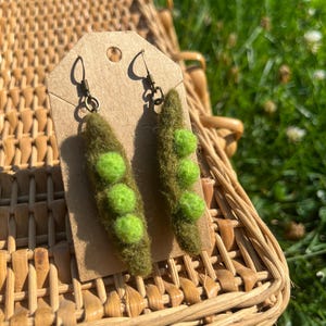 Puede incluir: Un par de pendientes colgantes con forma de vainas de guisantes de fieltro de color verde oliva, cada una con tres guisantes de fieltro de color verde lima brillante. Los pendientes están sujetos a una tarjeta de cartón marrón con ganchos de metal oscuro.