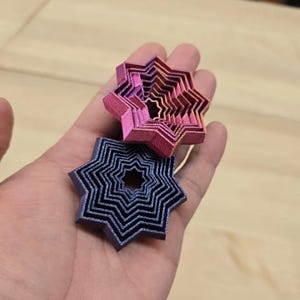 May include: Two layered star-shaped objects in hand. The top star is pink and orange, while the bottom star is blue and black. The objects have a layered design, creating a 3D effect. The background is a light brown wooden surface.