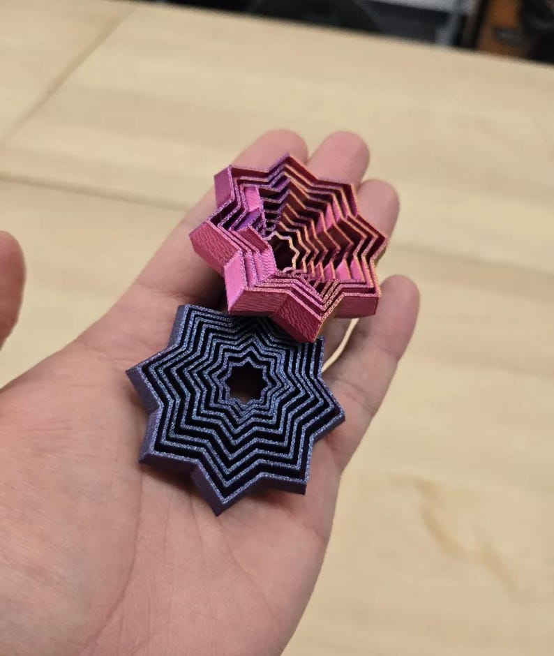 May include: Two layered star-shaped objects in hand. The top star is pink and orange, while the bottom star is blue and black. The objects have a layered design, creating a 3D effect. The background is a light brown wooden surface.
