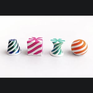 Puede incluir: Cuatro objetos decorativos en fila. El primero es un cono con rayas azules y verdes, el segundo es un cubo con rayas rosas y blancas, el tercero es una forma curva con rayas verdes y azules, y el último es una esfera con rayas naranjas y blancas.