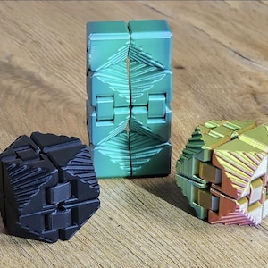 Puede incluir: Tres juguetes geométricos. Un juguete alto y verde articulado está de pie. También hay un juguete negro en forma de cubo y un juguete facetado multicolor. Los juguetes están sobre una superficie de madera.